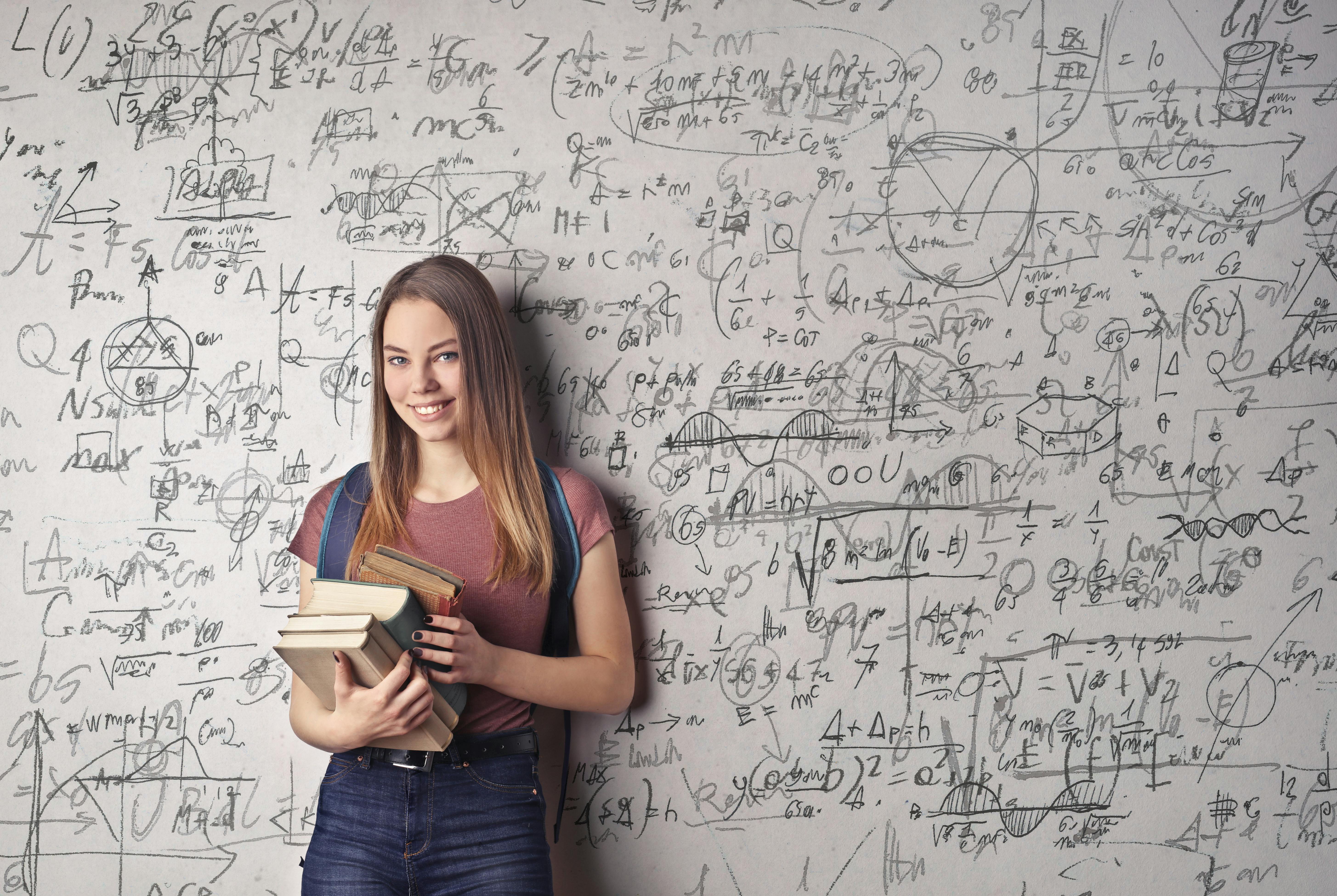 Frau mit drei Büchern in der Hand vor einem Whiteboard mit einer vielzahl an komplexen Formeln.